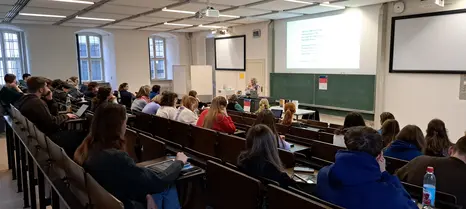 Das Foto zeigt den großen Hörsaal im Erdgeschoss des Gebäudes "An der Universität 5." Studierende sitzen auf stufig angeordneten Sitzbänken und hören der Vortragenden, Cris Miller, eine weiße Frau mit grauen, kinnlangen Haaren zu. Das Foto ist aus der rechten oberen Ecke des Hörsaals aufgenommen.