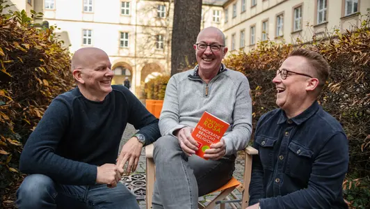 Thomas Weißer, Stefan Huber und Konstantin Lindner mit dem reisenden Regiestuhl auf dem Campus der Universität Bamberg. 