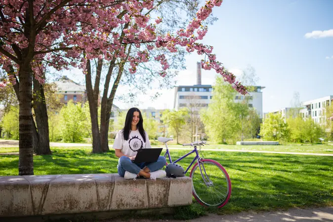Studentin sitzt mit einem Laptop vor einem blühendem Baum im ERBA-Park vor einem Universitätsgebäude