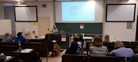 Das Foto zeigt den Hörsaal im Erdgeschoss des Gebäudes "An der Universität 5." Es ist von der Rückwand des Raumes aus aufgenommen, leicht nach rechts versetzt. Studierende sitzen auf stufig angeordneten Holzbänken. Die Gastrednerin Prof. Cris Miller, eine weiße Frau mit grauen, kinnlangen Haaren steht vor dem Rednerpult. Hinter ihr sind eine Tagel und auf der Wand die Zeilen eines Gedichts von Emily Dickinson zu sehen.